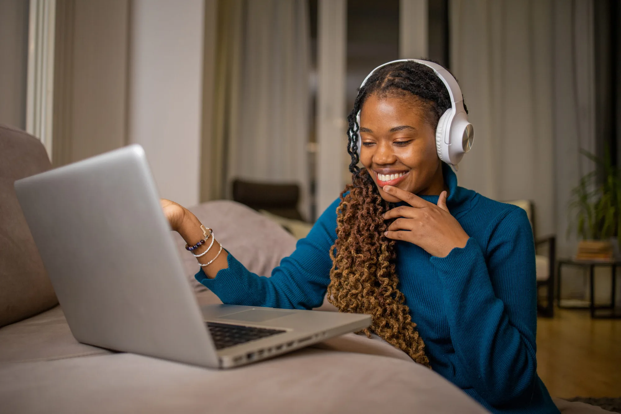 A smiling woman wearing headphones talks to someone on her laptop from her sofa.