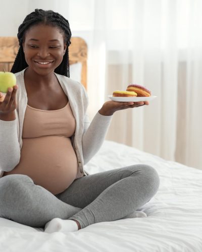 Pregnancy Diet. Smiling Pregnant Woman Choosing Green Apple Instead Of Donuts, Enjoying Organic Vitamin Fruits And Healthy Nutrition, Joyful African American Expectant Mother Sitting On Bed At Home